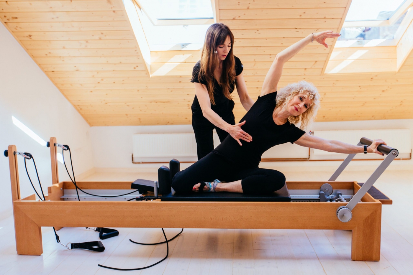 Sporty blond curly senior woman doing pilates exercises in gym with help from female physical therapist or instructor.